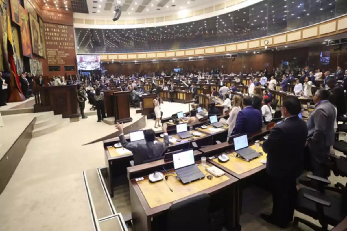 Overview of the interior of a parliament with numerous legislators sitting and standing at their desks, laptops on the tables, and an atmosphere of political debate in a spacious and well-lit chamber.