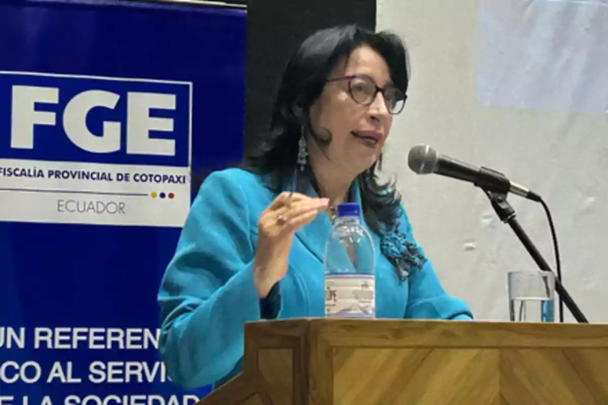 Dark-haired woman with glasses speaking at a podium with a microphone, wearing a blue blazer, next to a sign for the Provincial Prosecutor's Office of Cotopaxi in Ecuador. Dark-haired woman with glasses speaking at a podium with a microphone, wearing a blue blazer, next to a sign for the Provincial Prosecutor's Office of Cotopaxi in Ecuador.