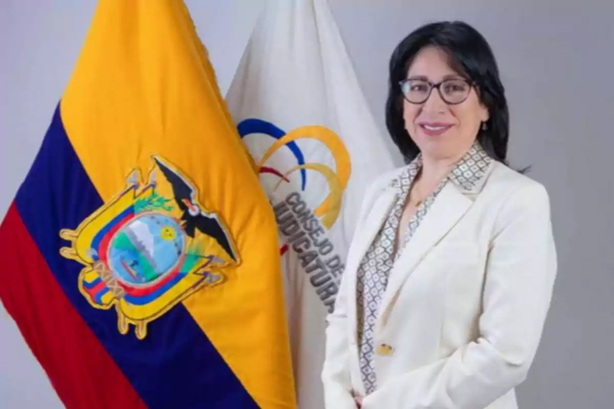 Woman wearing glasses and a white blazer posing next to the Ecuadorian flag and an institutional flag of the Judiciary Council