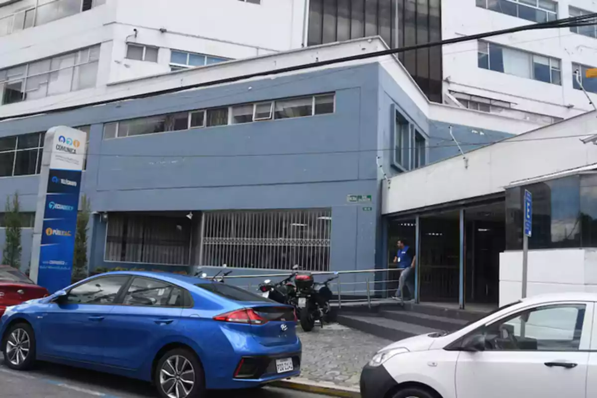 Facade of a blue office building with several cars and motorcycles parked in front and a person coming out through the access ramp