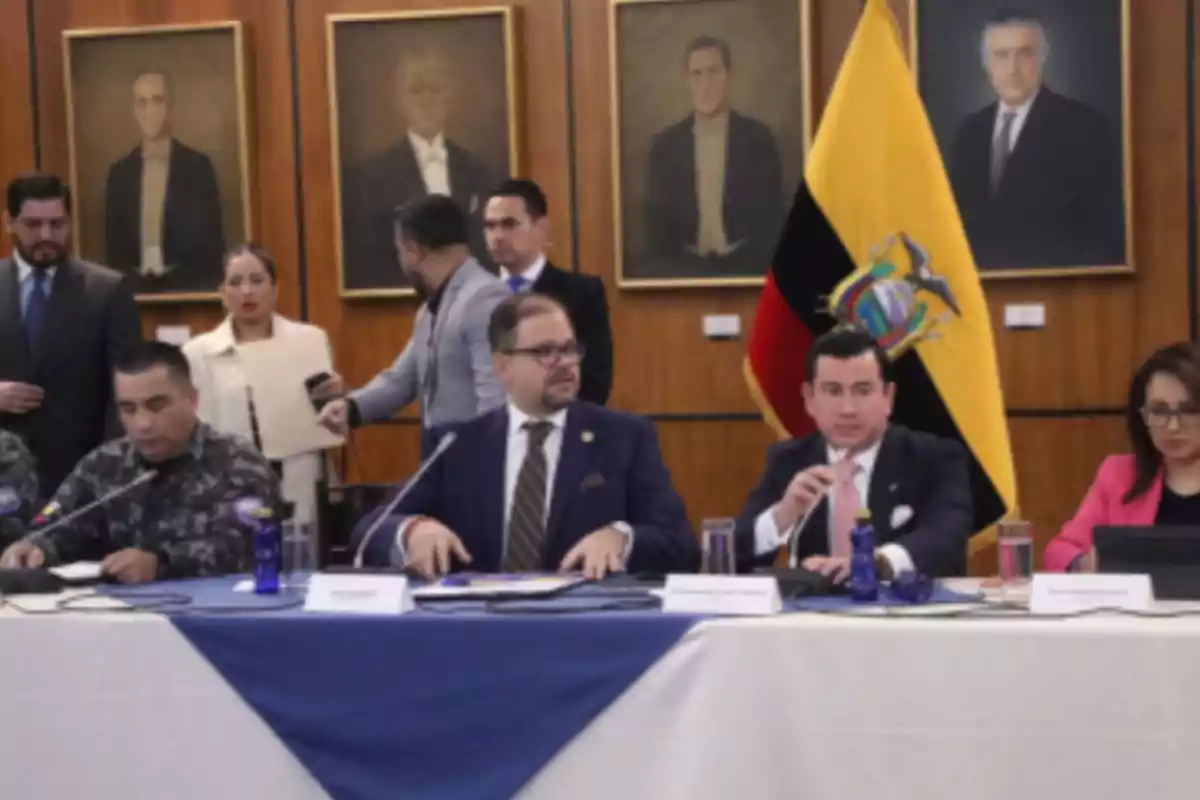 Varias personas sentadas en una mesa de reunión con la bandera de Ecuador al fondo y retratos en la pared Varias personas sentadas en una mesa de reunión con la bandera de Ecuador al fondo y retratos en la pared
