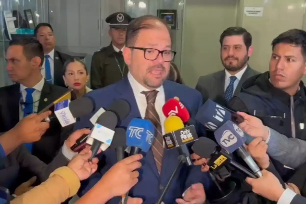 Un hombre con gafas y traje azul da declaraciones a la prensa rodeado de varios micrófonos y periodistas en un ambiente formal