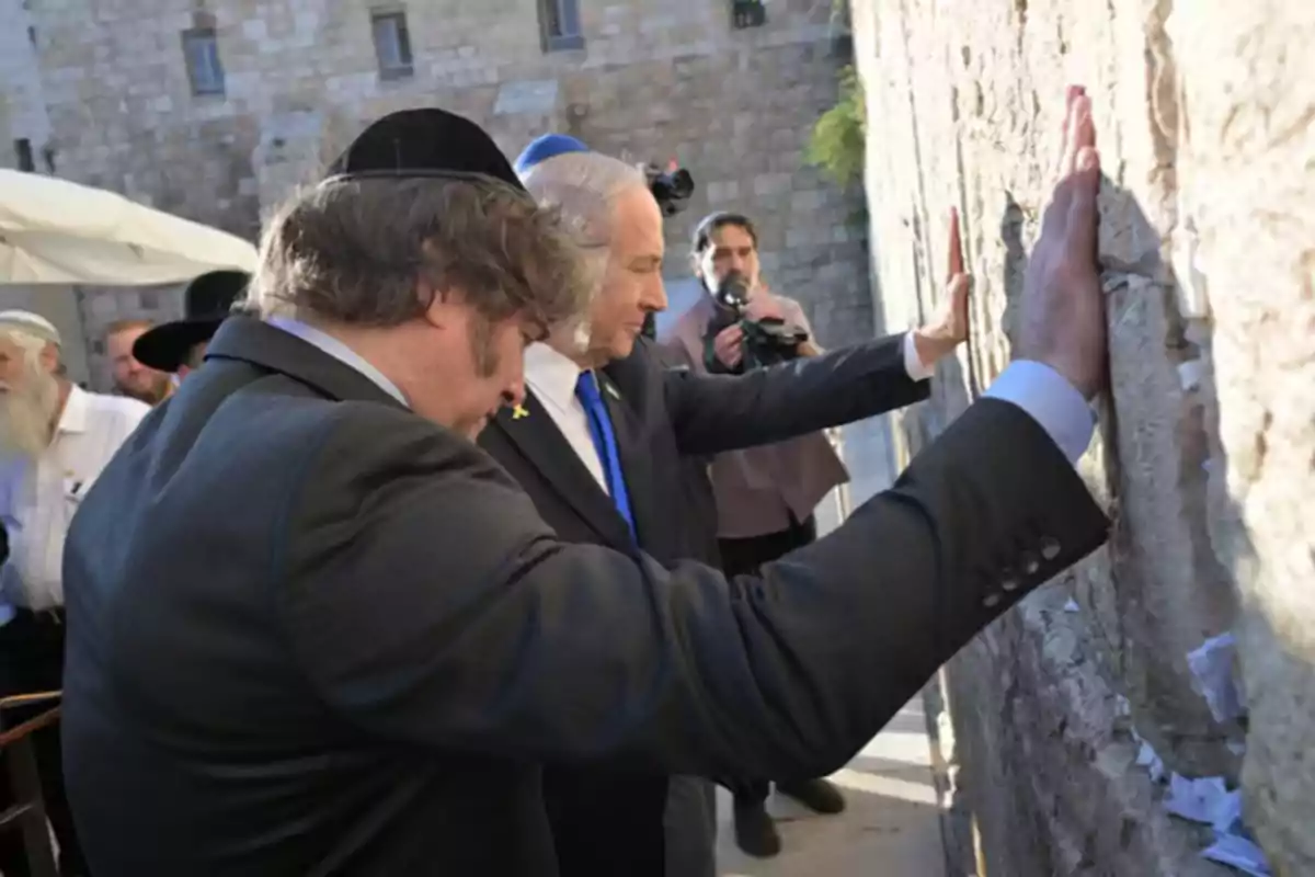 Dos hombres vestidos de traje rezan con las manos apoyadas en el Muro de los Lamentos mientras otras personas observan y un camarógrafo graba la escena. Dos hombres vestidos de traje rezan con las manos apoyadas en el Muro de los Lamentos mientras otras personas observan y un camarógrafo graba la escena.