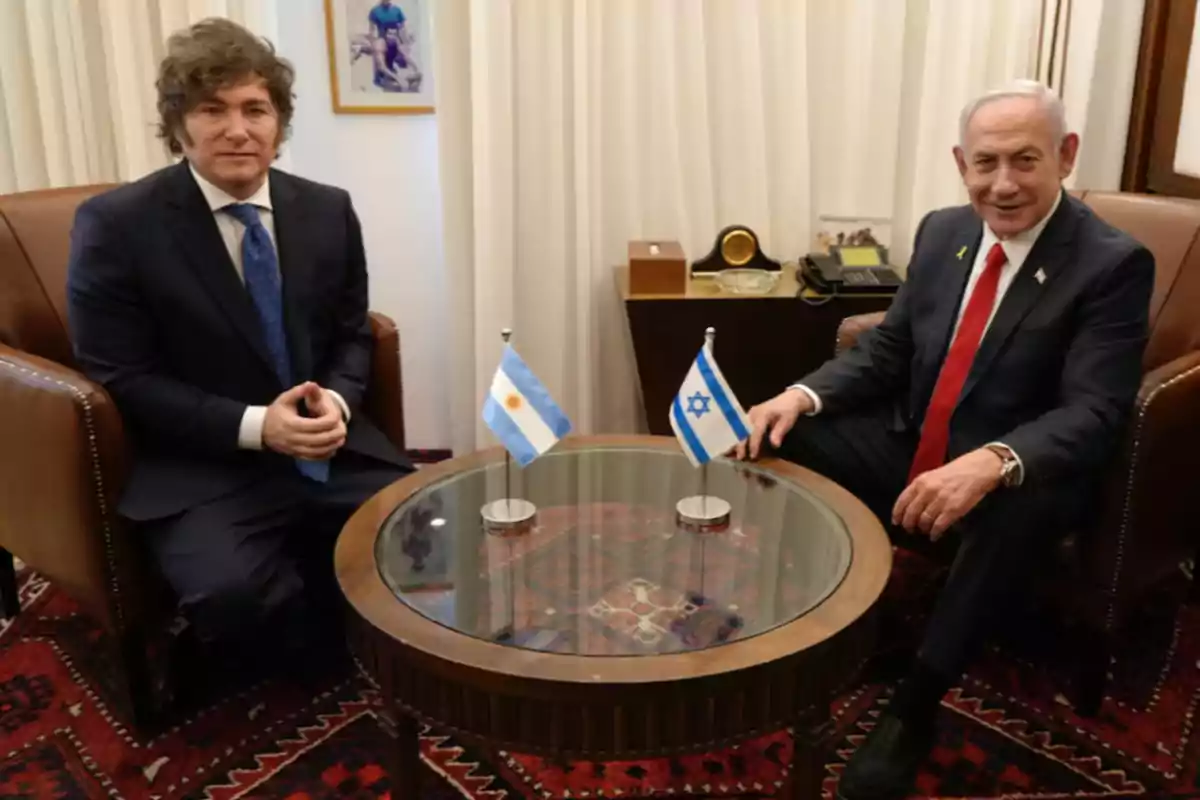 Dos hombres con traje sentados en sillones alrededor de una mesa de vidrio con las banderas de Argentina e Israel en el centro