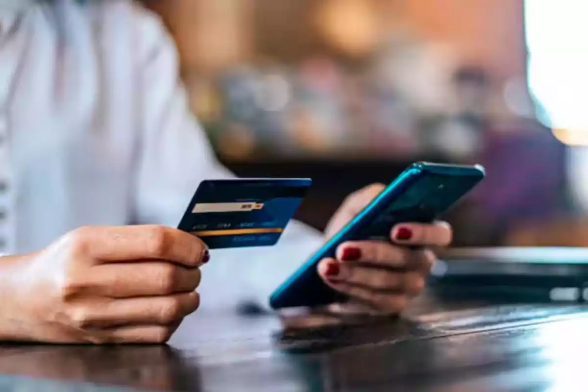 Persona sosteniendo una tarjeta bancaria en una mano y un teléfono móvil en la otra sobre una mesa Persona sosteniendo una tarjeta bancaria en una mano y un teléfono móvil en la otra sobre una mesa