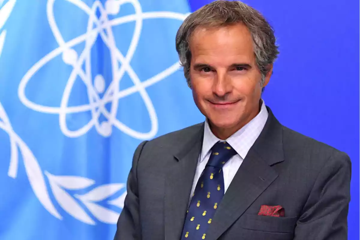 Hombre de cabello canoso con traje y corbata posando frente a una bandera azul con un símbolo atómico blanco