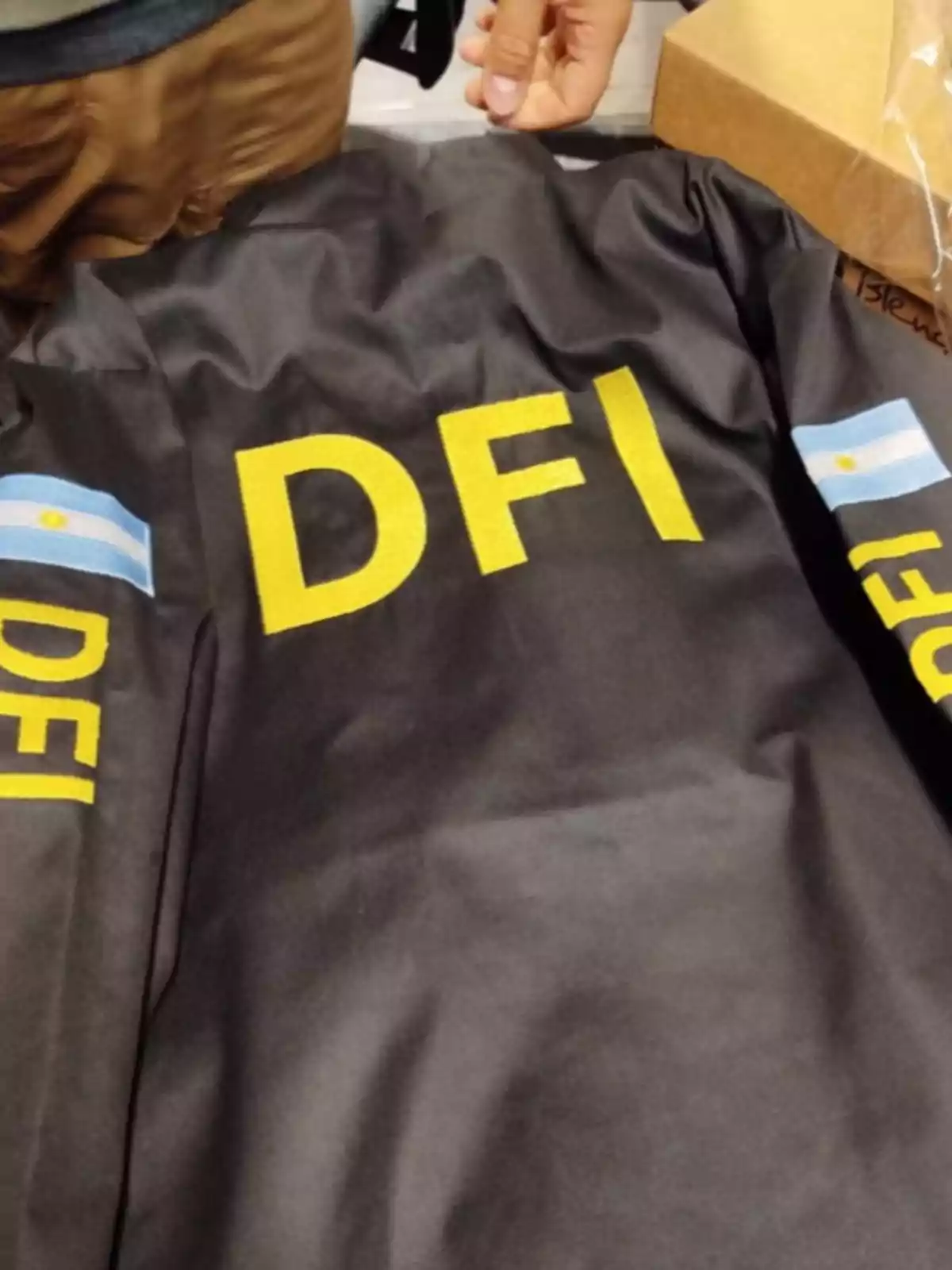 Chamarra negra con las letras DFI en amarillo y banderas de Argentina en las mangas, sobre una mesa junto a una mano y una caja de cartón Chamarra negra con las letras DFI en amarillo y banderas de Argentina en las mangas, sobre una mesa junto a una mano y una caja de cartón