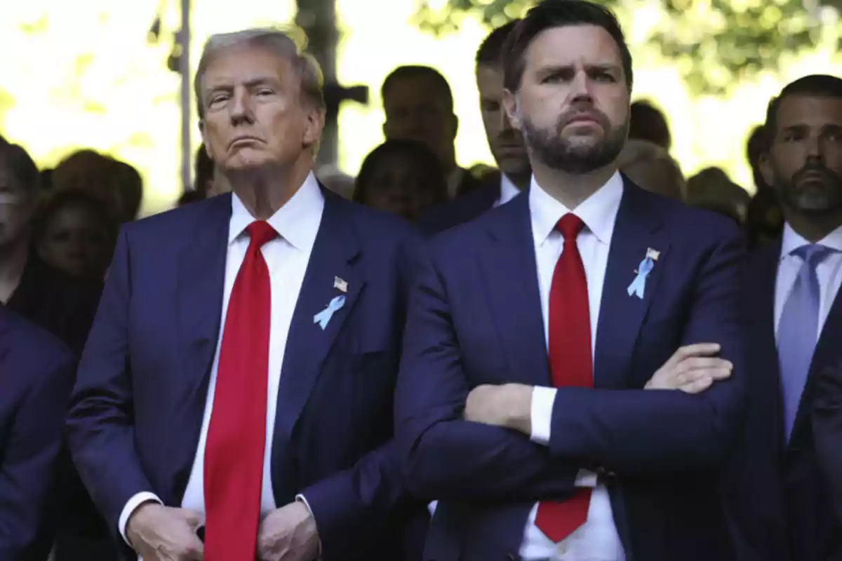 Dos hombres de traje azul y corbata roja con listones azules en la solapa están de pie en un evento solemne rodeados de otras personas