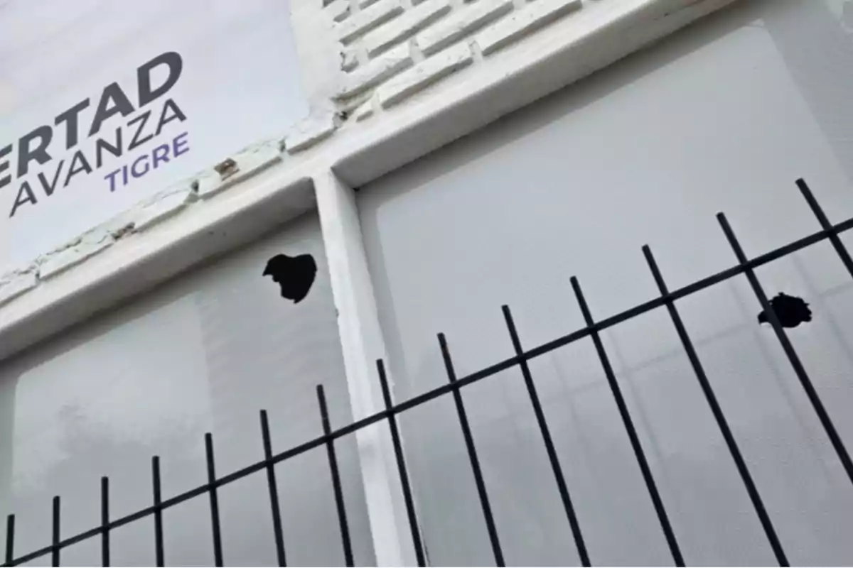 Broken window with two holes and a black grille in front; in the upper left corner, there is a sign with partial text. Broken window with two holes and a black grille in front; in the upper left corner, there is a sign with partial text.