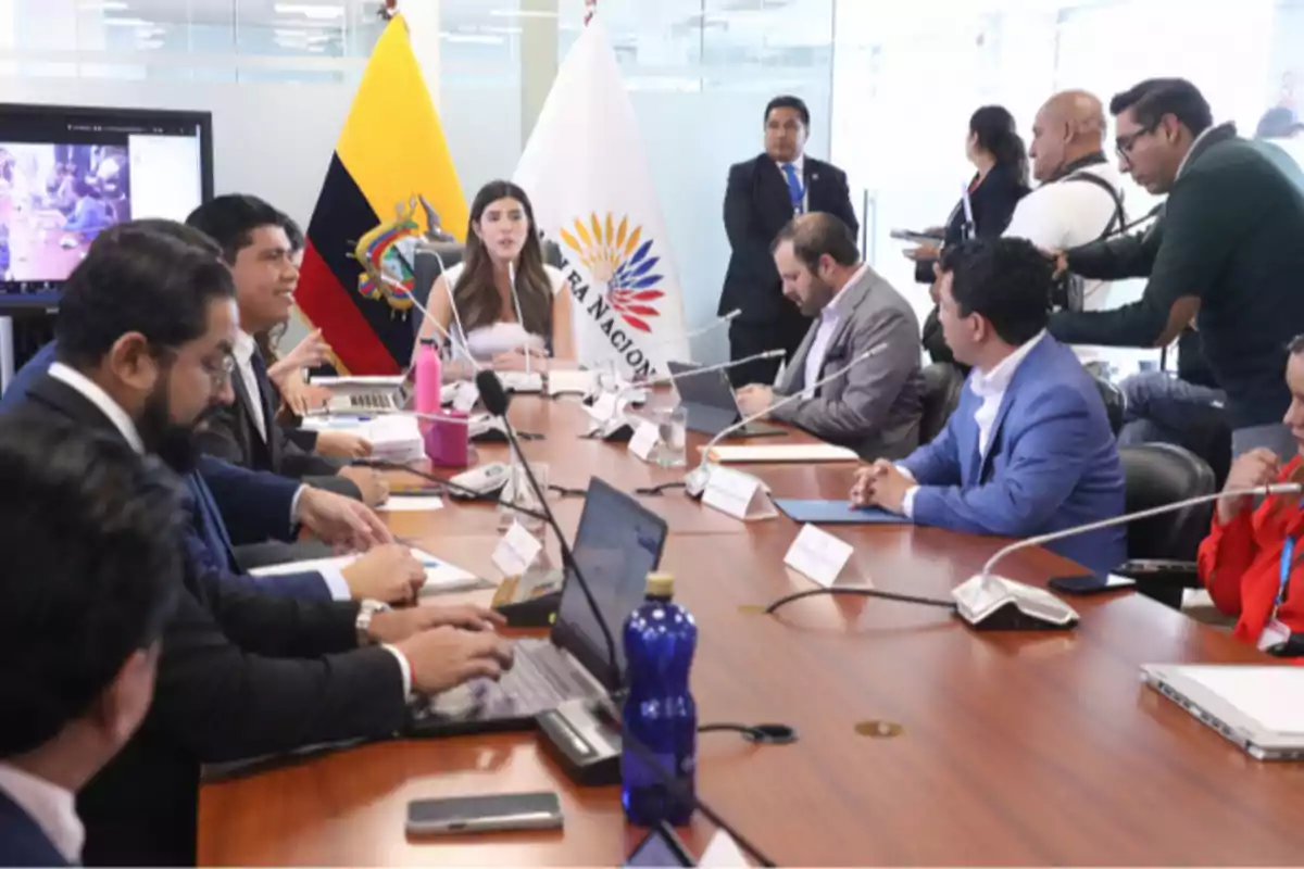 People gathered in a boardroom with Ecuadorian and National Assembly flags in the background, while some work on laptops and others take notes, in a formal work environment.