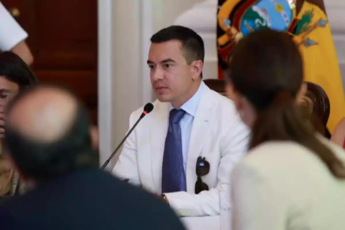 Man in a white suit and blue tie speaking at a formal meeting with a microphone in front of him and the Ecuadorian flag in the background