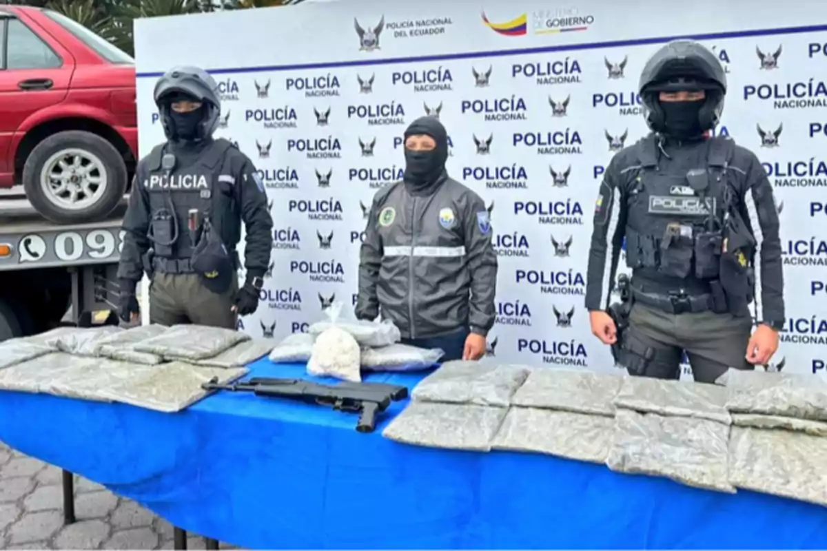 Three police officers pose in front of a table with seized drug packages and a weapon, with an institutional National Police of Ecuador backdrop and a red vehicle in the background.