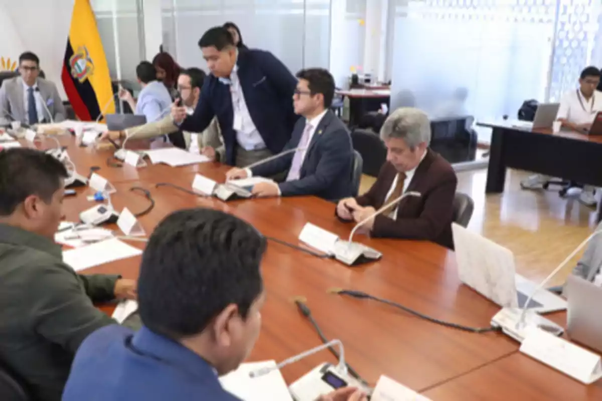 Varias personas sentadas alrededor de una mesa de reuniones en una oficina con una bandera de Ecuador al fondo
