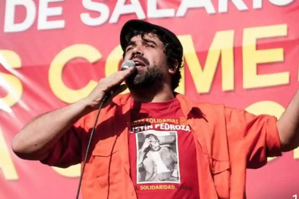 Hombre con micrófono hablando frente a un cartel rojo con letras amarillas.