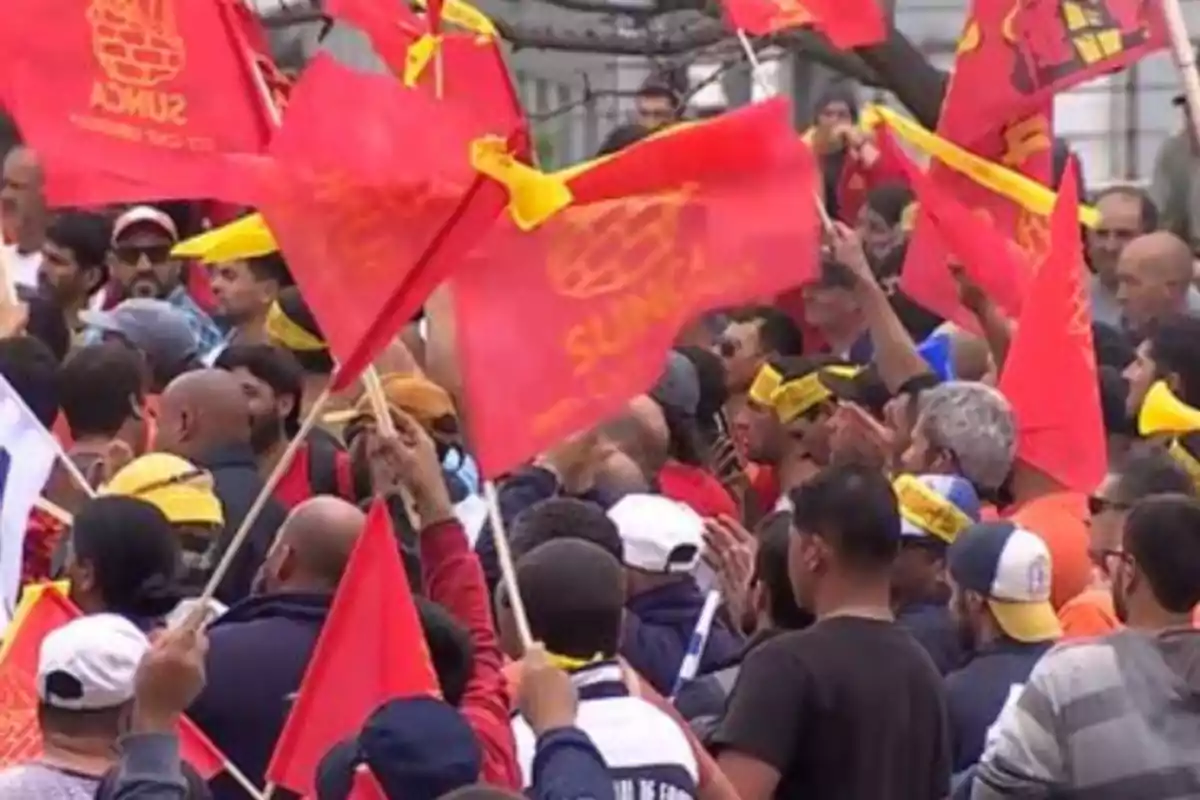 Un grupo de personas se reúne en una manifestación, ondeando banderas rojas con inscripciones amarillas.