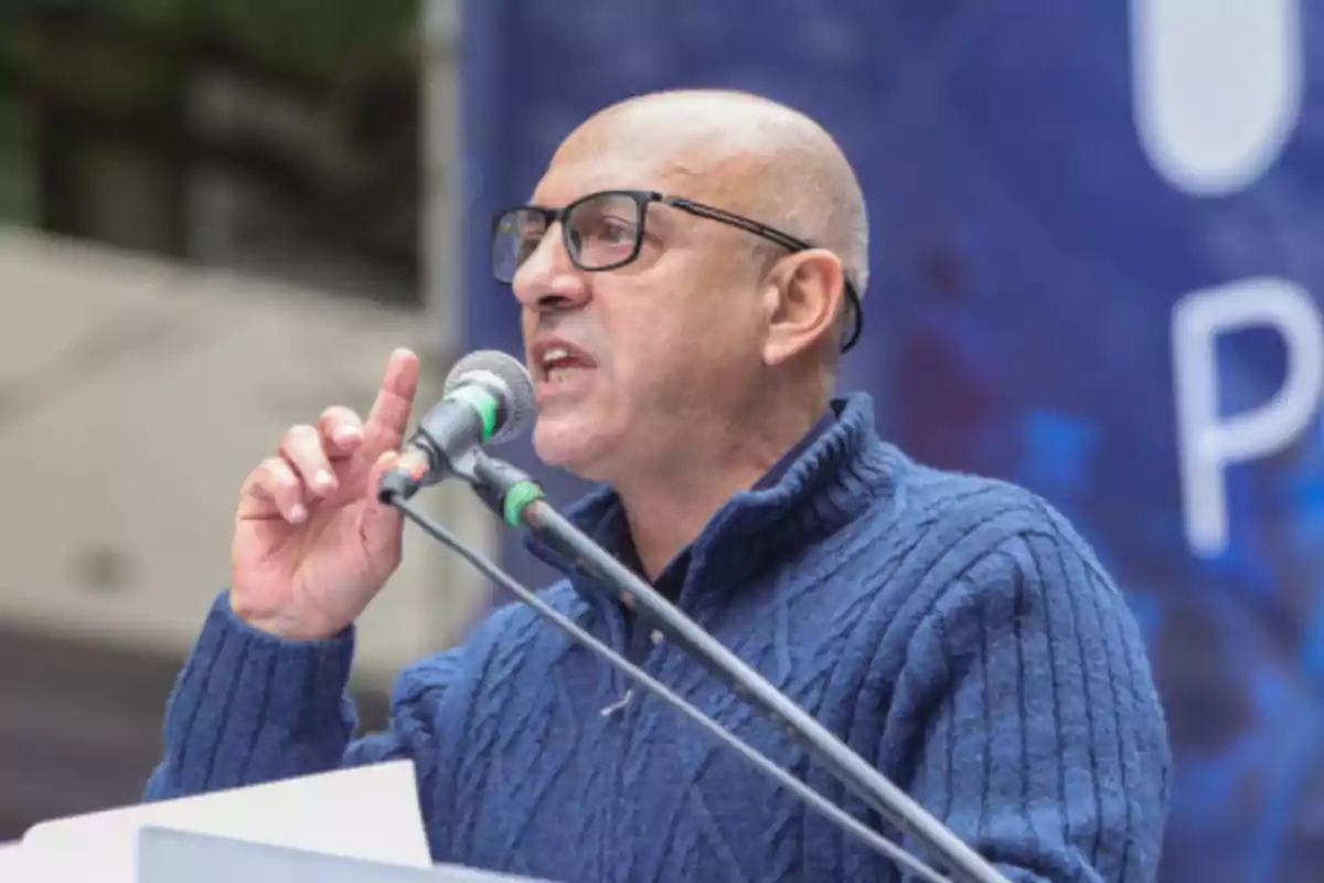Bald man with glasses and a blue sweater speaking into a microphone during a public event