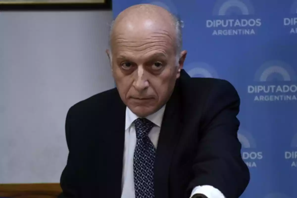 Un hombre calvo con traje oscuro y corbata está frente a un fondo azul que dice Un hombre calvo con traje oscuro y corbata está frente a un fondo azul que dice