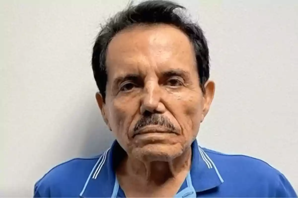 Un hombre mayor con bigote y cabello oscuro viste una camisa azul y está frente a un fondo claro.