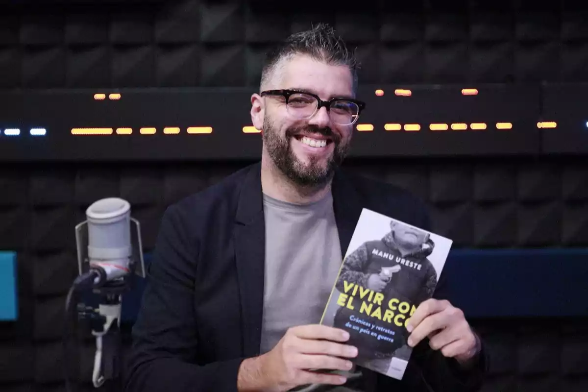 Un hombre con gafas y barba sonríe mientras sostiene un libro titulado 