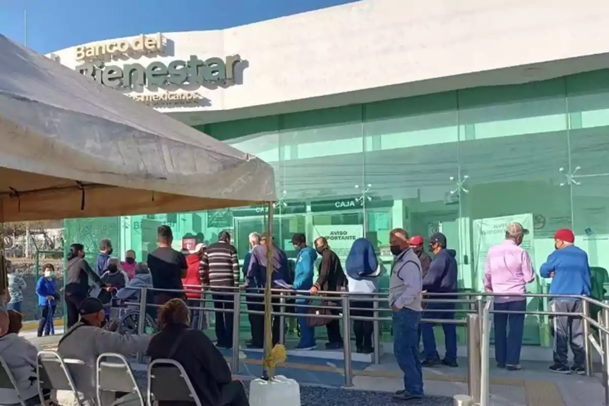 Personas haciendo fila afuera de una sucursal del Banco del Bienestar en un día soleado.