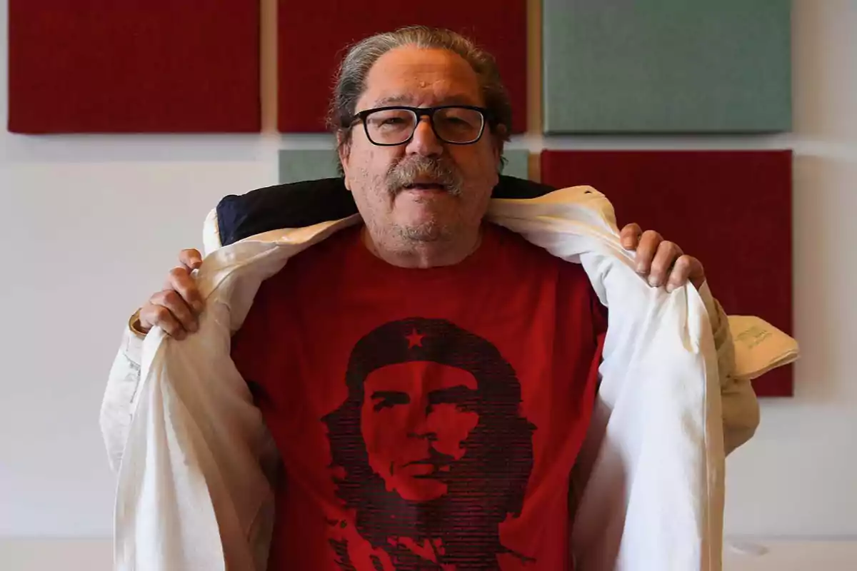 Un hombre con gafas y bigote muestra una camiseta roja con la imagen del Che Guevara mientras sostiene abierta una chaqueta blanca.