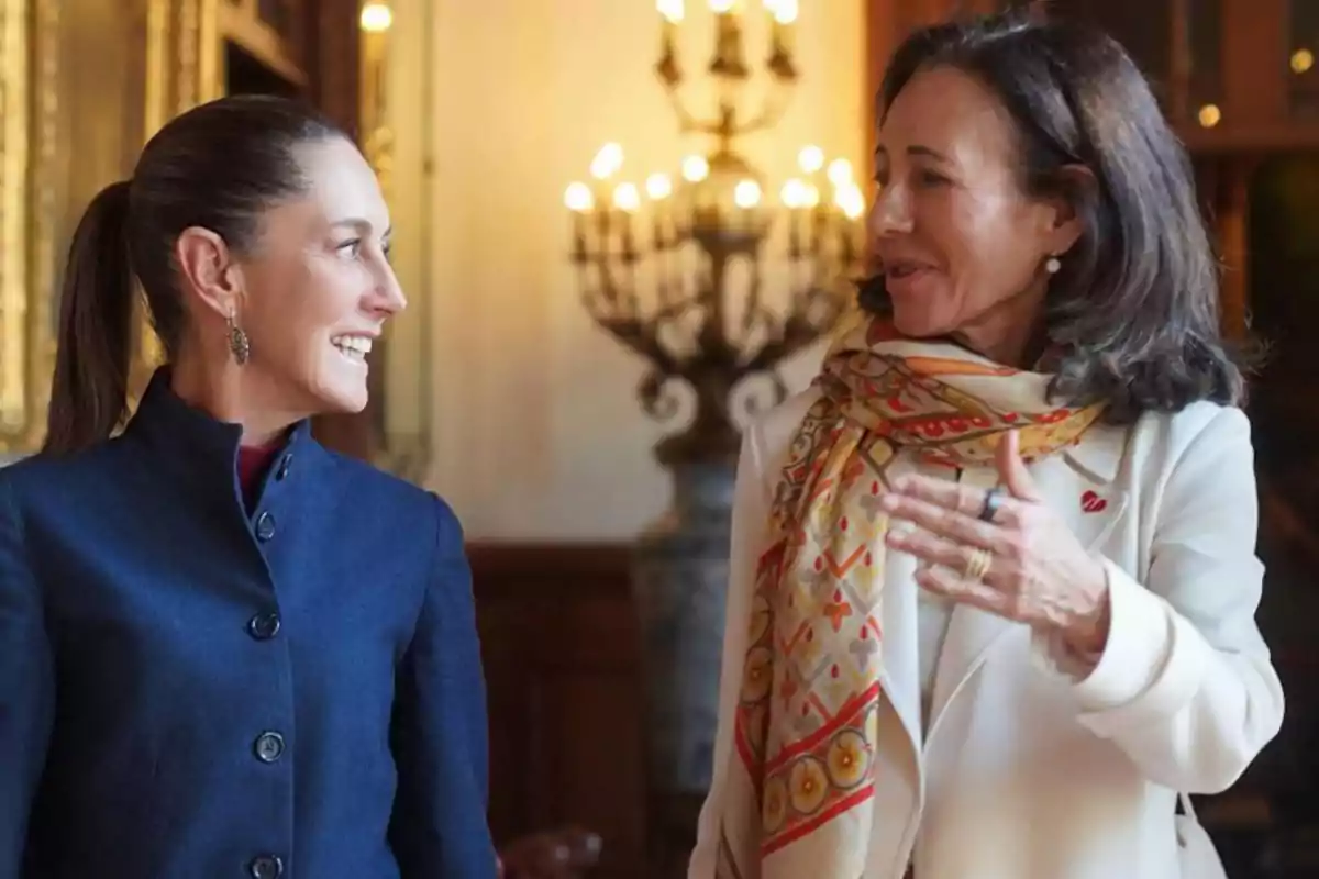 Dos mujeres conversando y sonriendo en un ambiente elegante con candelabros de fondo.