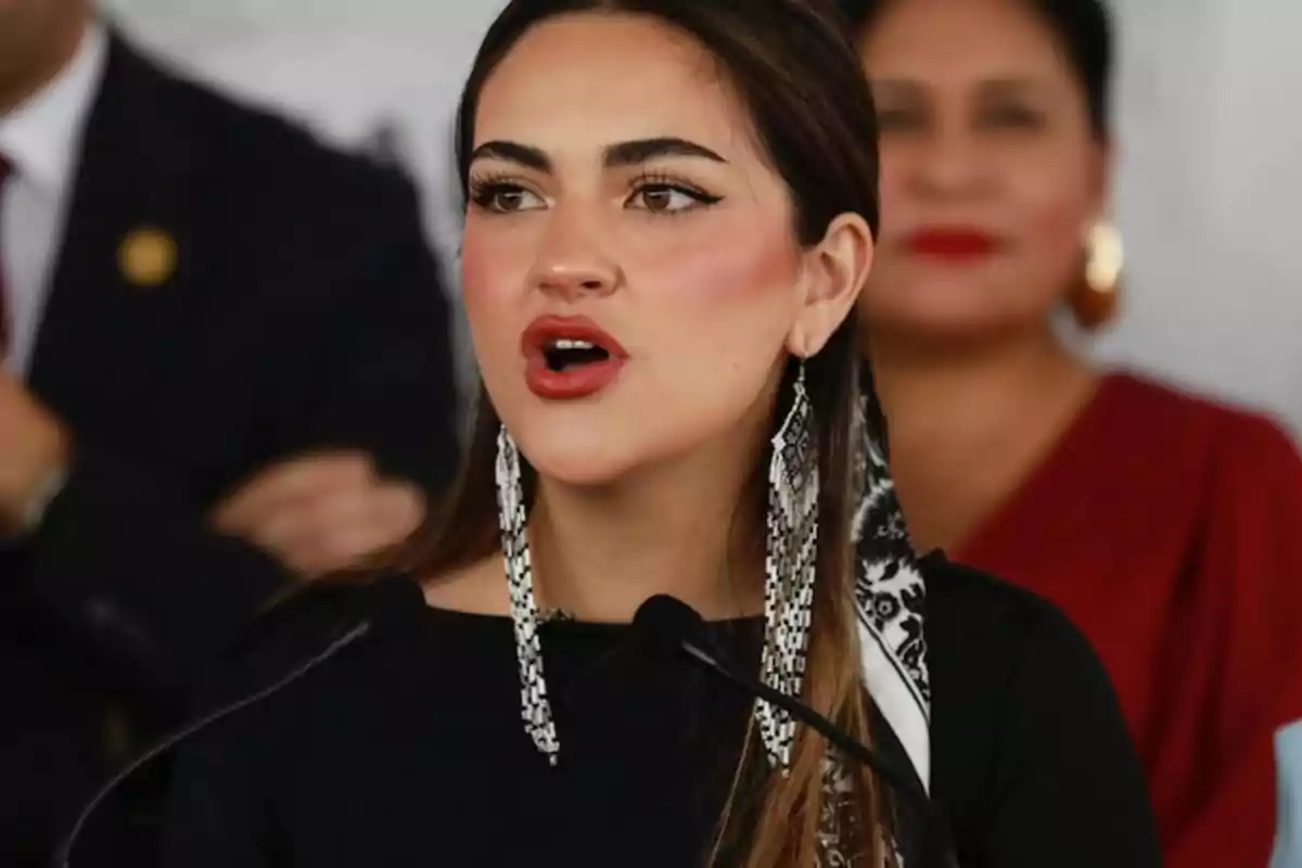 A woman with long earrings and a scarf in her hair speaks in front of a microphone with blurred people in the background.