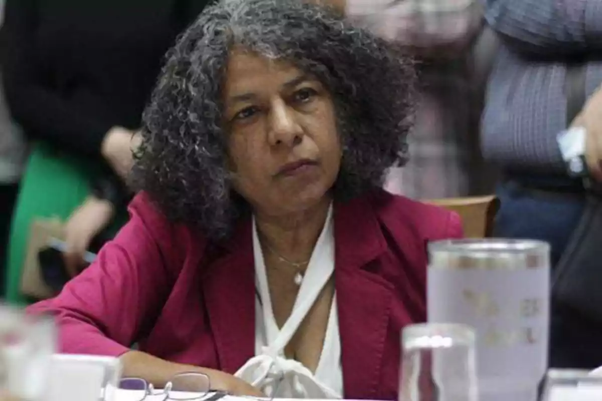 A person with curly gray hair is sitting at a table, wearing a red jacket and a white blouse, with a thoughtful expression.