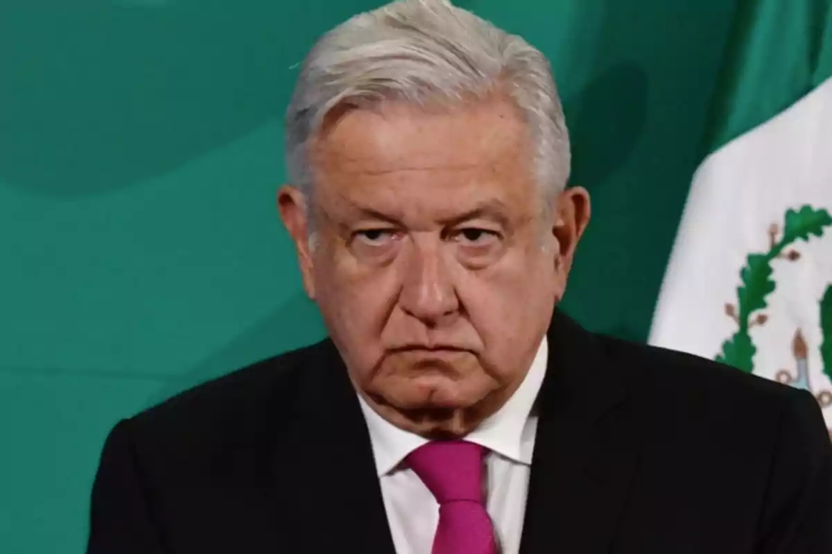 Un hombre de cabello canoso y expresión seria con una corbata rosa y un fondo verde con parte de una bandera visible.