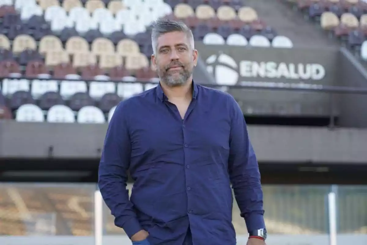 Hombre de cabello canoso y barba con camisa azul de pie en un estadio con gradas vacías al fondo y un letrero que dice ENSALUD