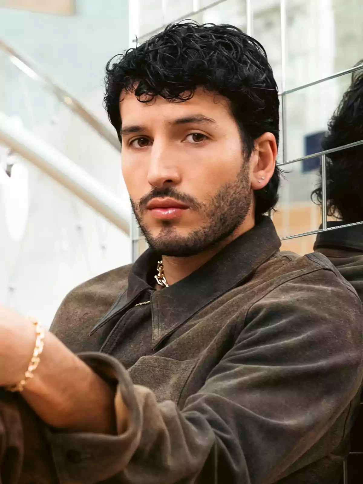 A man with curly hair and a beard, wearing a brown jacket, is sitting in front of a mirror. A man with curly hair and a beard, wearing a brown jacket, is sitting in front of a mirror.