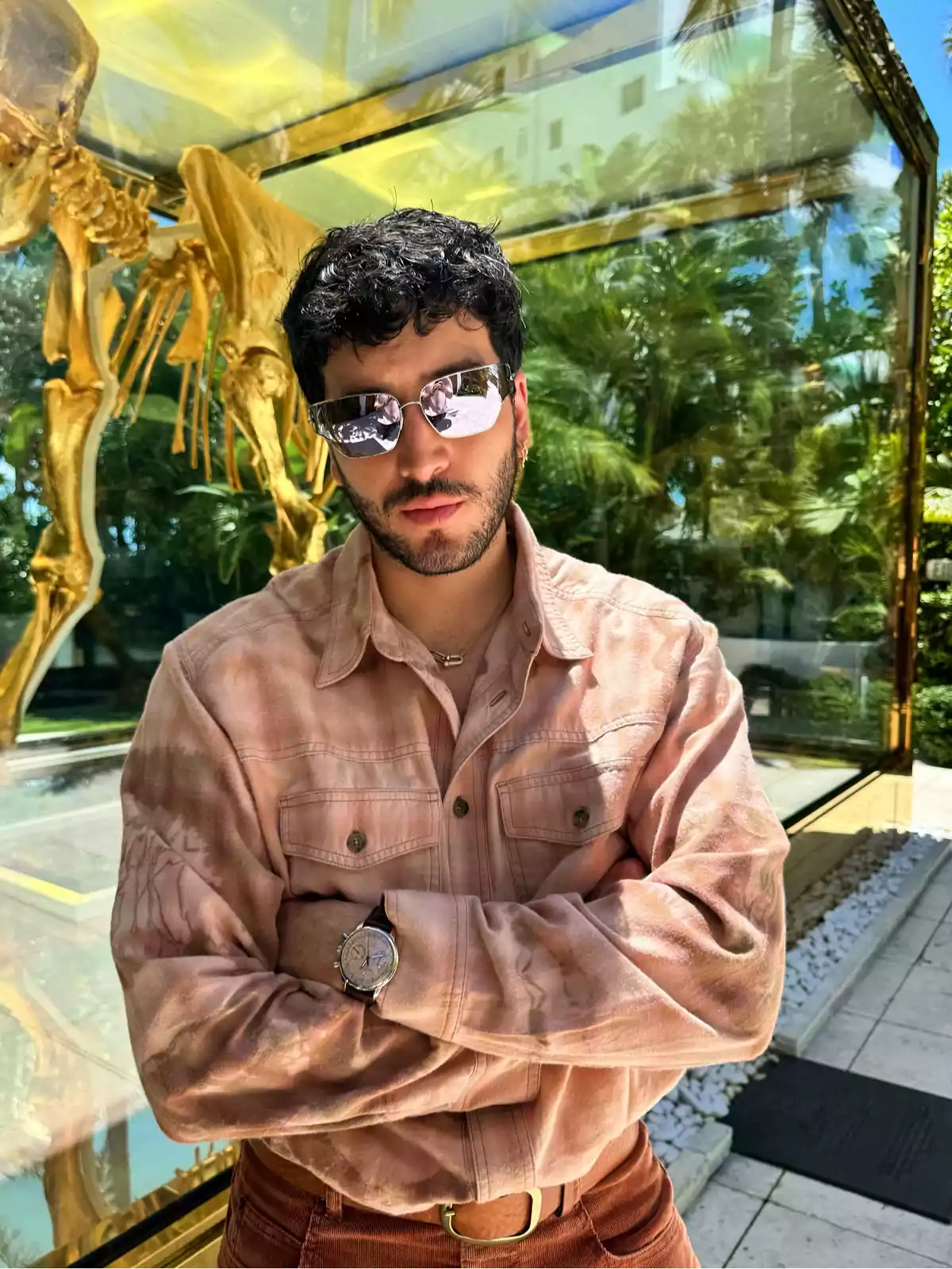 A man with sunglasses and a pink jersey poses with his arms crossed in front of a glass structure containing a golden skeleton, surrounded by vegetation. A man with sunglasses and a pink jersey poses with his arms crossed in front of a glass structure containing a golden skeleton, surrounded by vegetation.