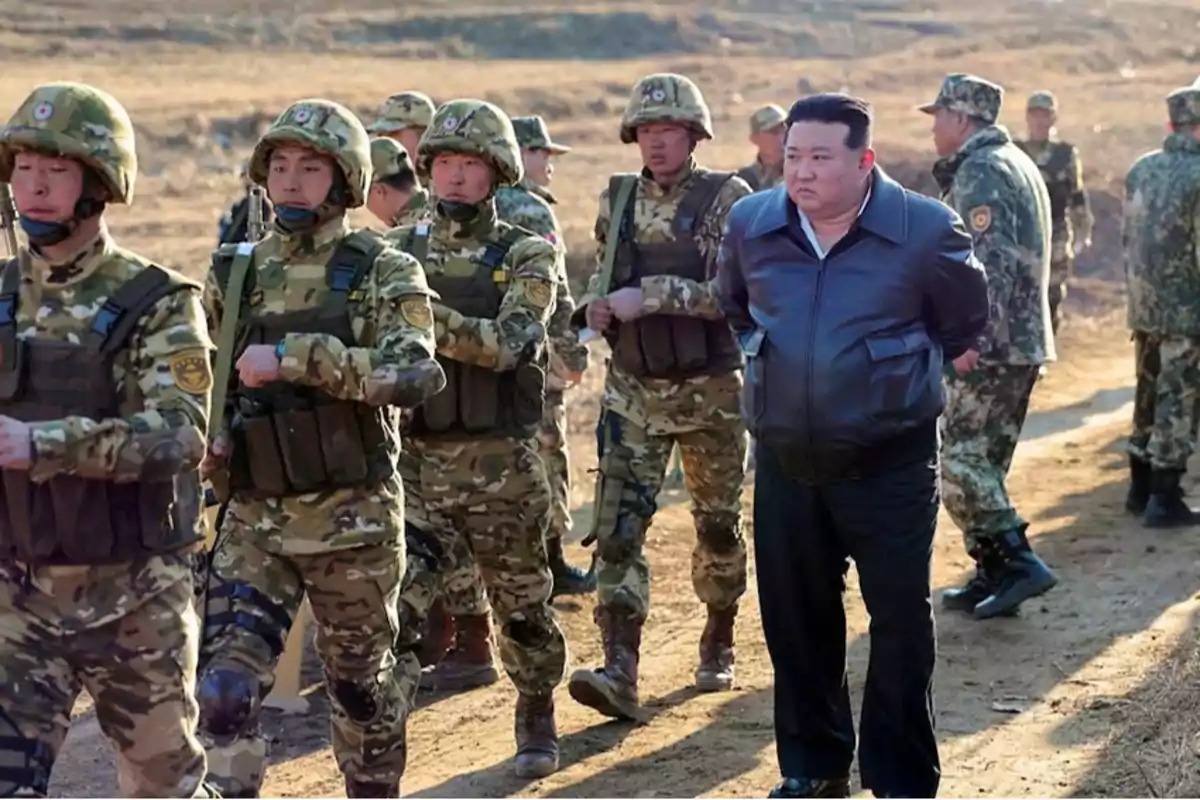 Un grupo de soldados en uniforme camuflado marcha junto a un hombre con chaqueta negra en un entorno al aire libre.