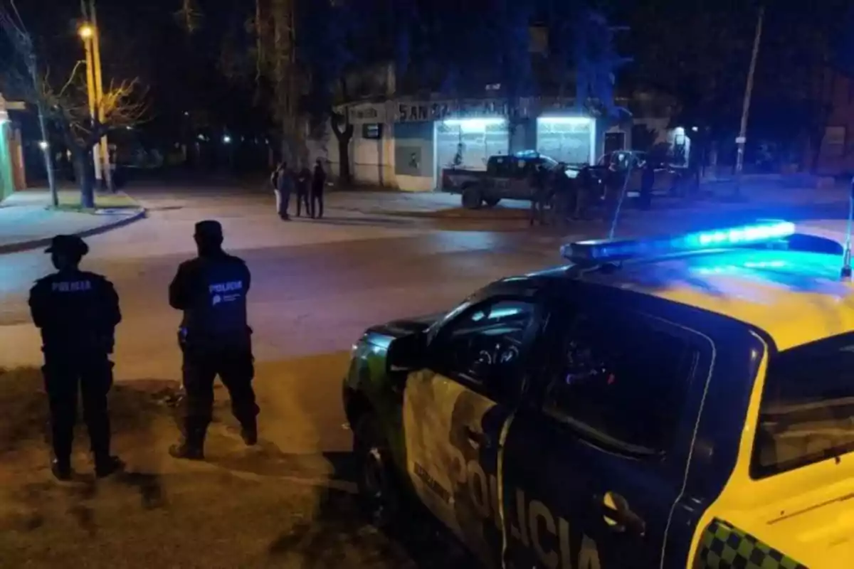 Policías de pie junto a un vehículo patrulla con luces encendidas en una calle por la noche.