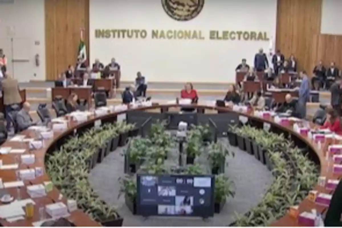 Reunión en una sala del Instituto Nacional Electoral con personas sentadas alrededor de una mesa circular y plantas en el centro.