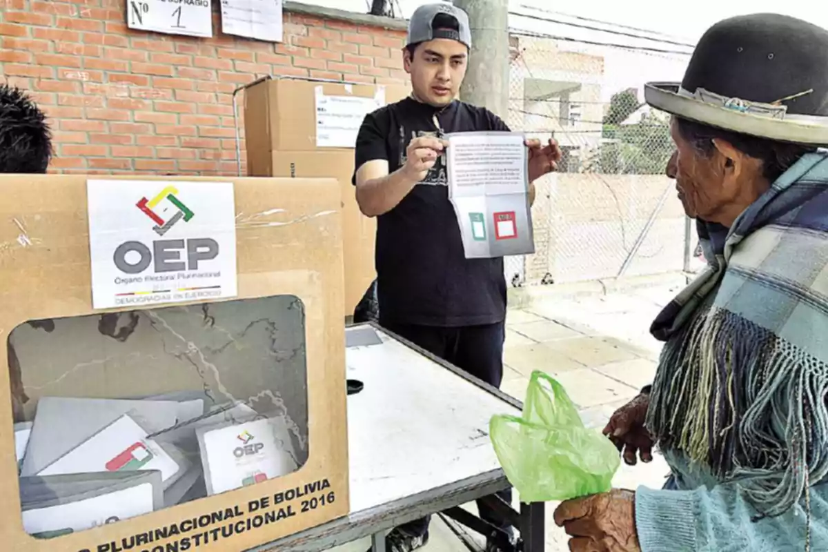 Un hombre muestra una papeleta de votación a una mujer mayor en un centro electoral de Bolivia durante un referéndum, con una urna de cartón del Órgano Electoral Plurinacional en primer plano. Un hombre muestra una papeleta de votación a una mujer mayor en un centro electoral de Bolivia durante un referéndum, con una urna de cartón del Órgano Electoral Plurinacional en primer plano.