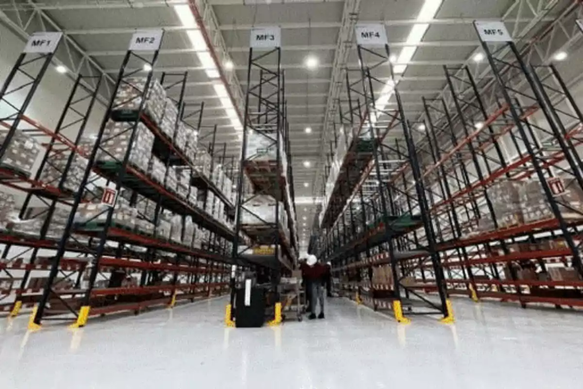 Interior de un almacén con estanterías altas llenas de cajas y una persona trabajando en el pasillo central.