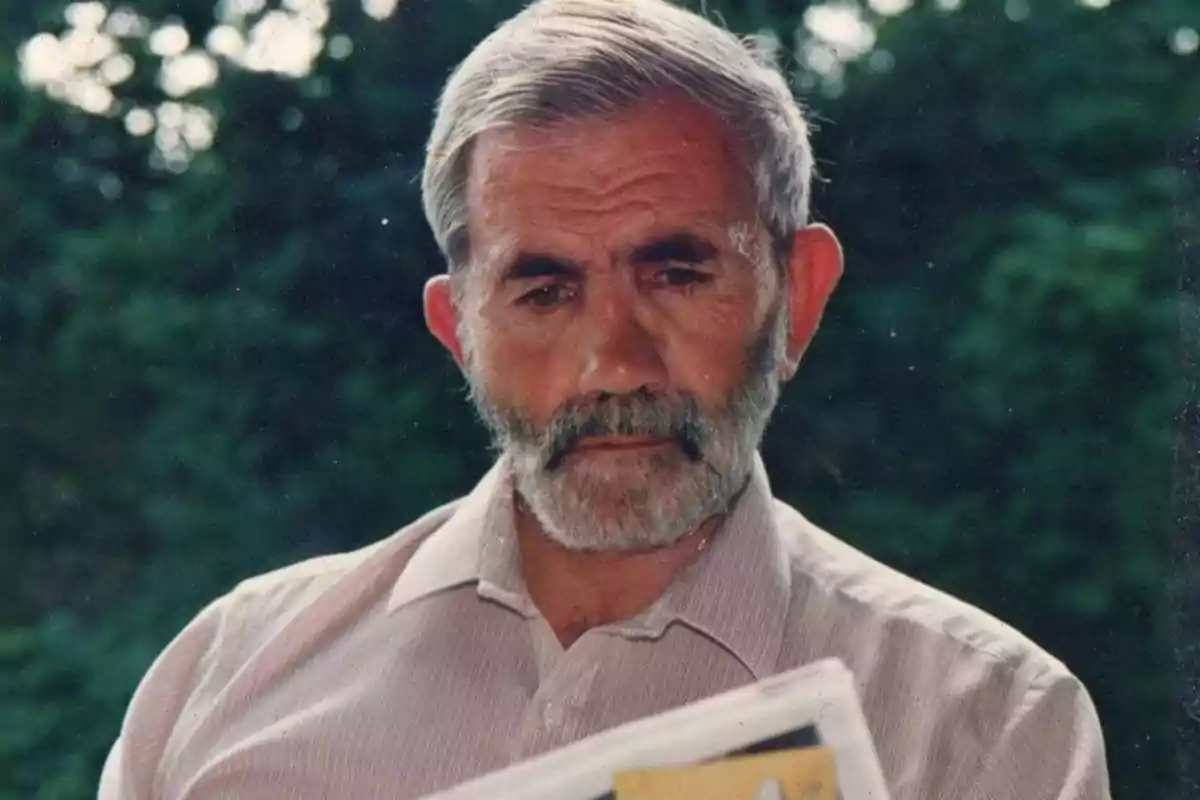 An elderly man with a gray beard and hair is reading a newspaper outdoors.