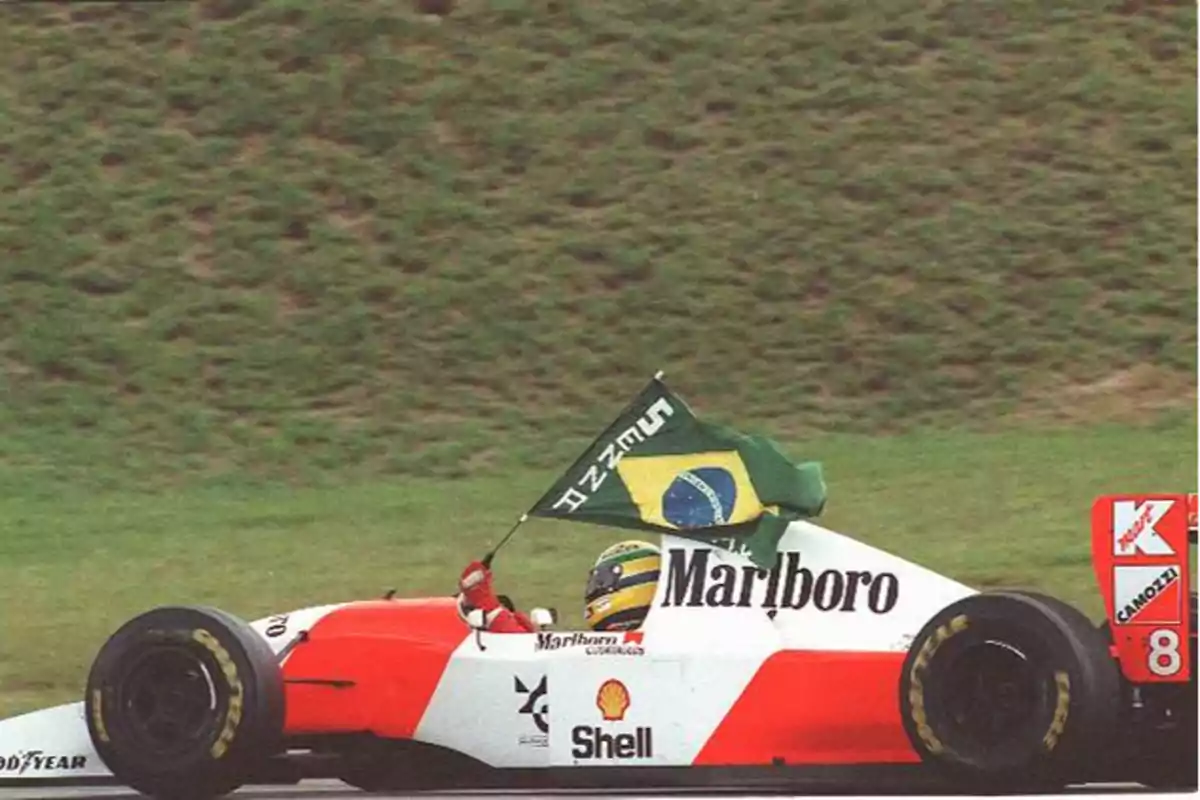 Un piloto de Fórmula 1 conduce un monoplaza con los colores rojo y blanco mientras sostiene una bandera de Brasil.