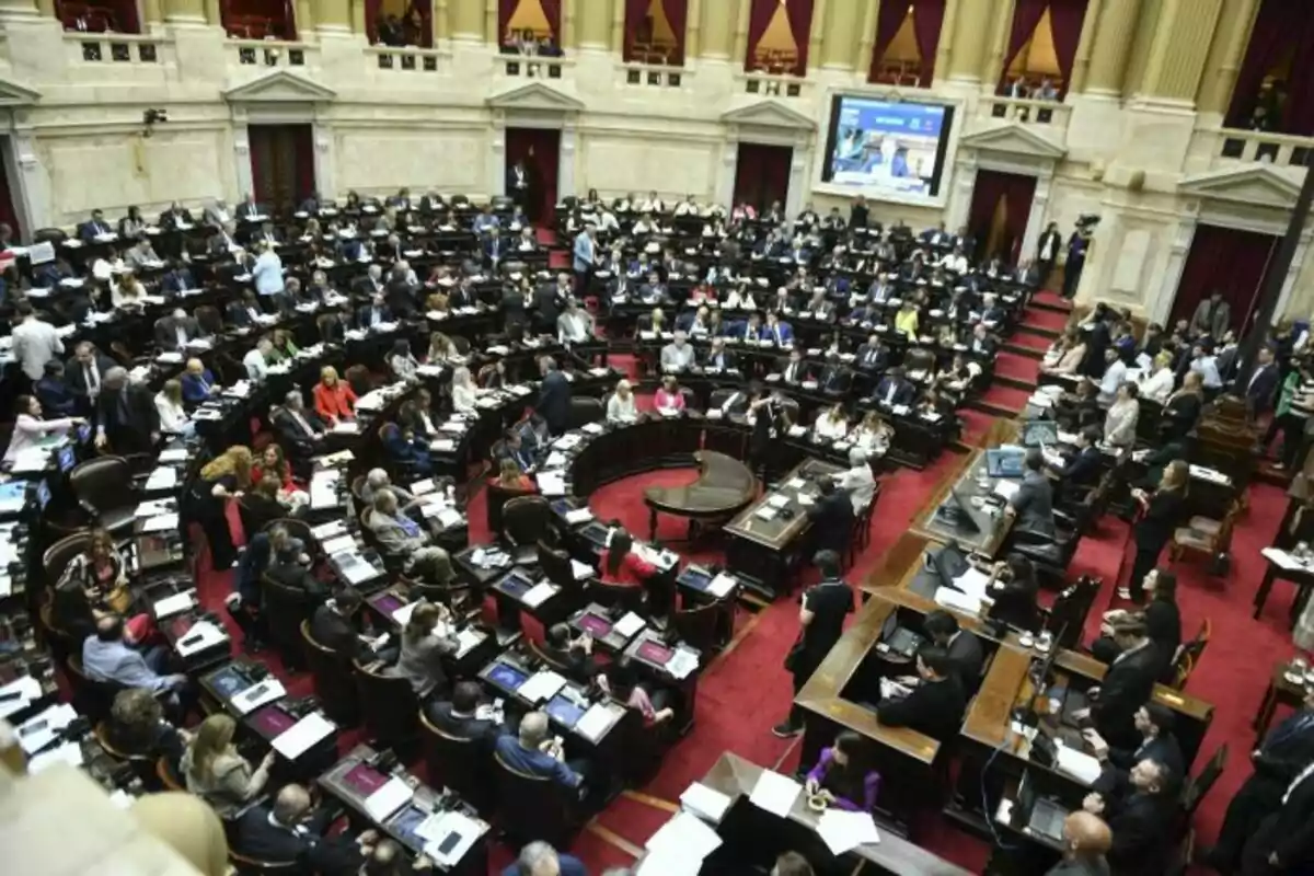 Una sesión parlamentaria en un recinto con numerosos legisladores sentados en sus escaños y un ambiente de debate activo.