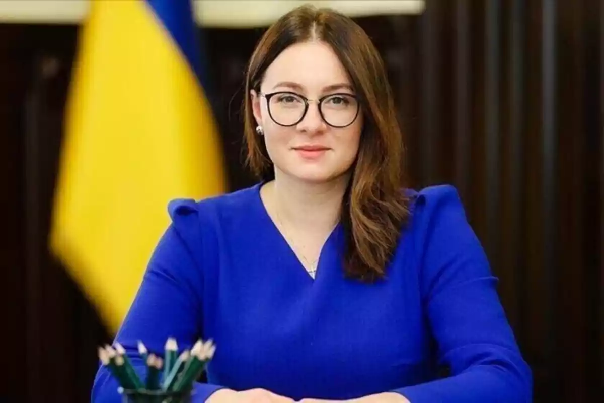 Una persona con gafas y cabello castaño viste de azul frente a una bandera. Una persona con gafas y cabello castaño viste de azul frente a una bandera.