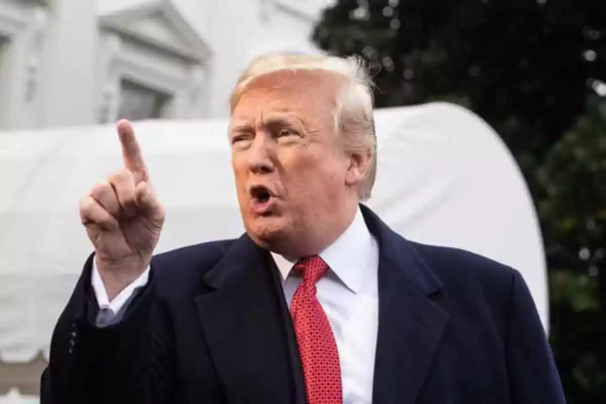 Blond-haired man in a dark suit and red tie raising his index finger while speaking in an outdoor setting