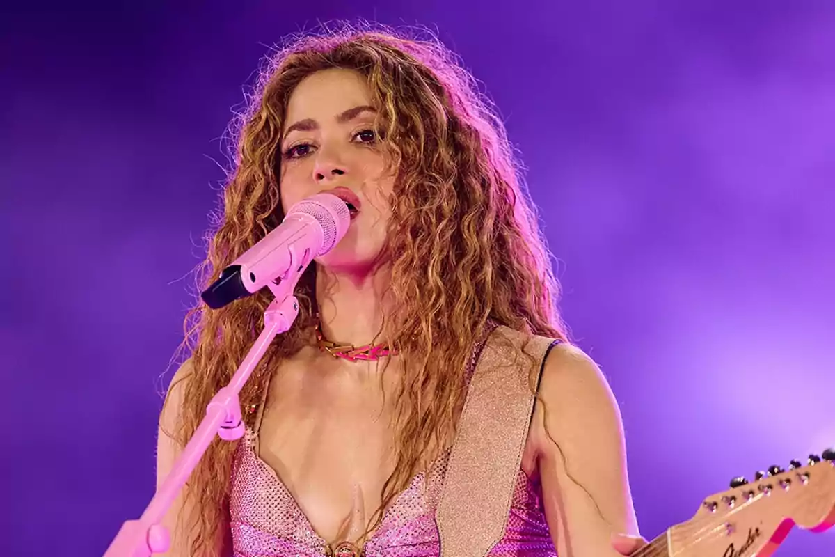 Una persona cantando en un escenario con un micrófono rosa y fondo púrpura.