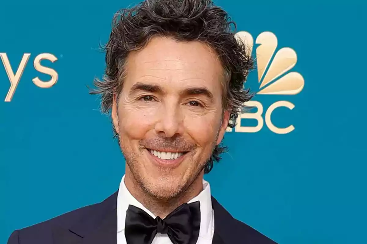 Hombre de cabello rizado y canoso con barba ligera usando esmoquin y pajarita negra sonriendo frente a un fondo azul con el logo de NBC Hombre de cabello rizado y canoso con barba ligera usando esmoquin y pajarita negra sonriendo frente a un fondo azul con el logo de NBC