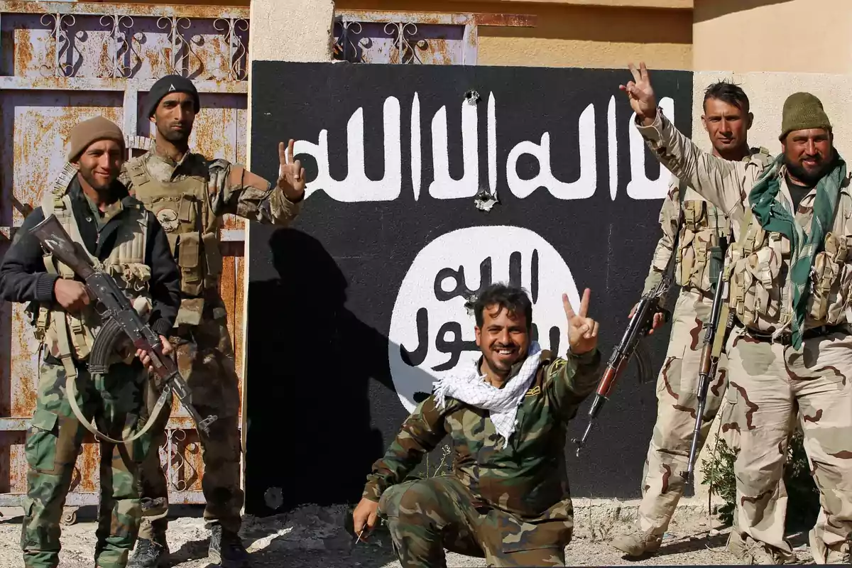 Un grupo de hombres armados en uniforme militar posando frente a una pared con un símbolo y texto en árabe. Un grupo de hombres armados en uniforme militar posando frente a una pared con un símbolo y texto en árabe.