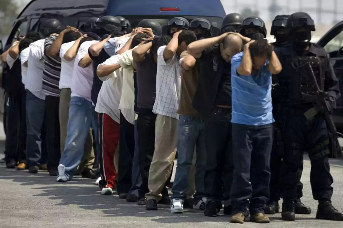 A group of people with their hands on their heads is guarded by heavily armed police officers in tactical gear next to a pickup truck. A group of people with their hands on their heads is guarded by heavily armed police officers in tactical gear next to a pickup truck.