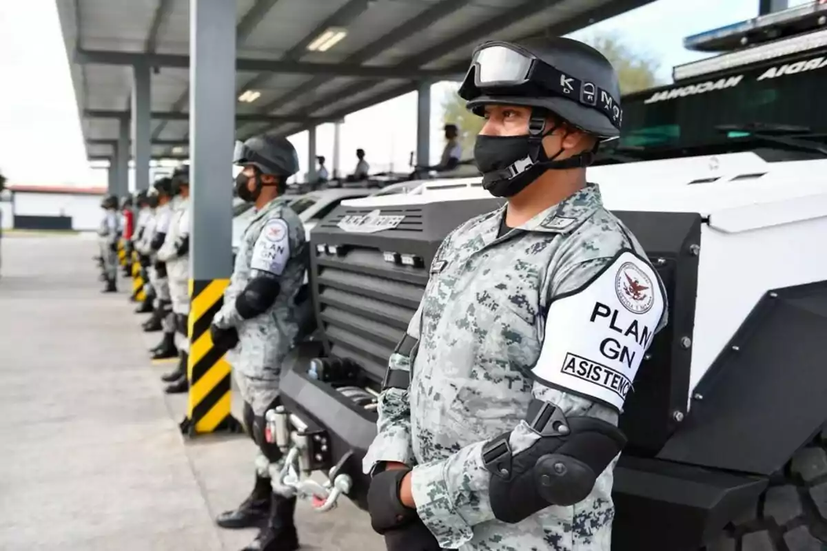 Members of the Mexican National Guard in tactical uniforms and protective gear are lined up next to armored vehicles under a covered structure. Members of the Mexican National Guard in tactical uniforms and protective gear are lined up next to armored vehicles under a covered structure.