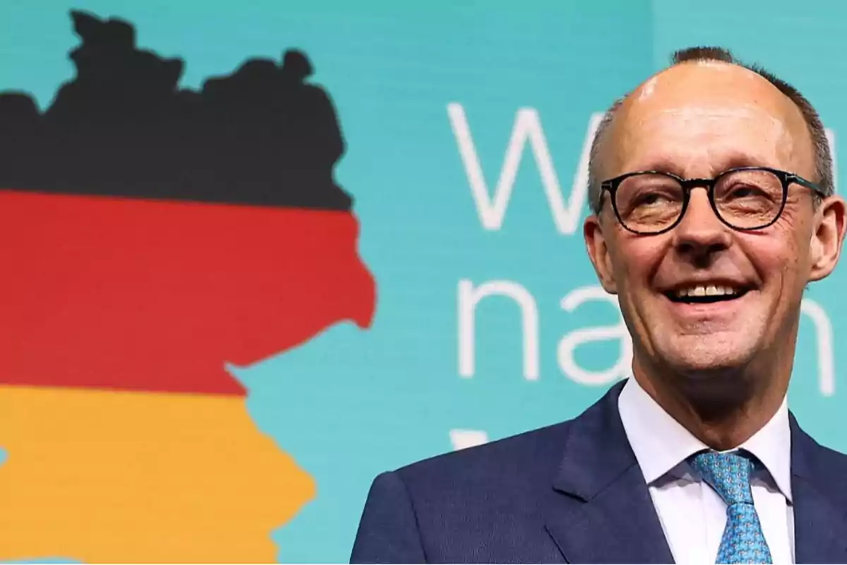 Un hombre con gafas y traje sonríe frente a un fondo azul que muestra un mapa de Alemania en colores negro, rojo y amarillo.