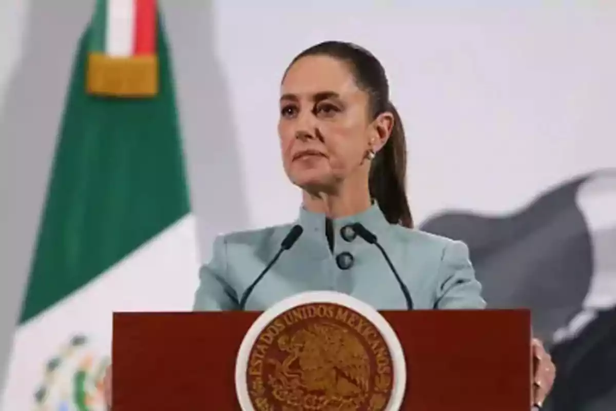 Una persona hablando en un podio con el escudo de México y una bandera mexicana de fondo.