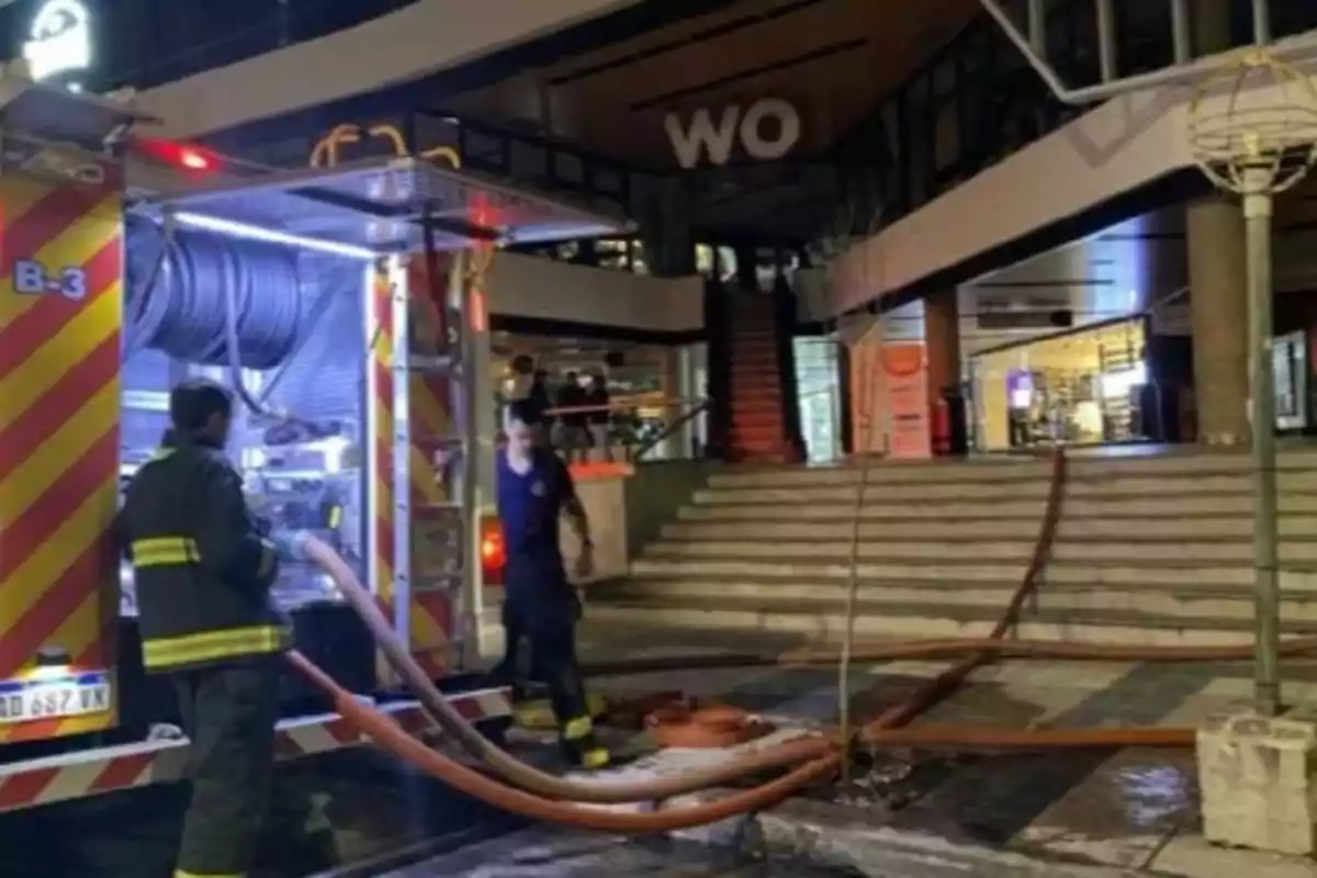 Bomberos trabajando con una manguera conectada a un camión frente a un edificio comercial por la noche Bomberos trabajando con una manguera conectada a un camión frente a un edificio comercial por la noche